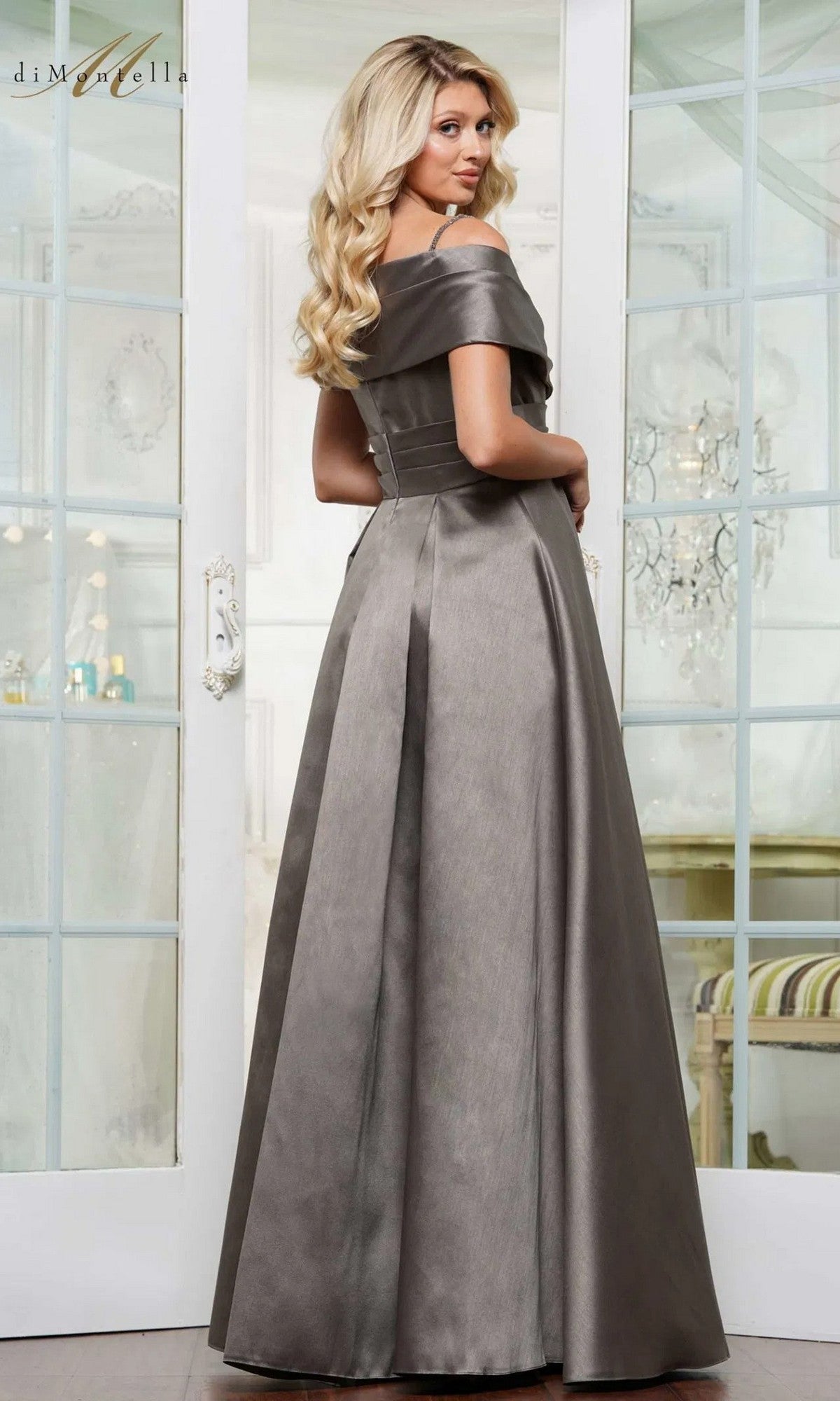 A woman with long blonde hair wears the Rina Di Montella RD3221, a floor-length silver-gray formal dress with a pleated skirt, standing indoors before glass doors.